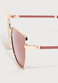 Sončna očala z zlato kovinsko ogrodje kvadratne oblike, roza gradacijskimi lečami, tankimi burgundskimi kraki in logotipom CK na tečaju.