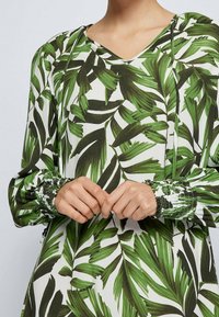 Langärmlige Bluse mit einem weißen Hintergrund und großen grünen Blattmustern. Geraffte Manschetten mit einem strukturierten Design in Grüntönen.