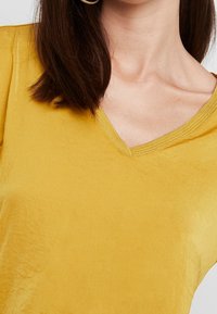 Vrouw met schouderlang bruin haar, die een mosterdgele V-hals t-shirt draagt, weergegeven van de nek tot halverwege de romp.