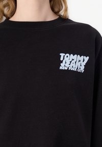Černá mikina s kulatým výstřihem a dlouhými rukávy. Na přední straně se nachází bílé stylizované logo "TOMMY JEANS NEW YORK CITY". Měkká textura.
