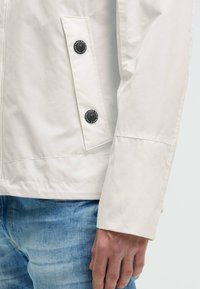 Giacca beige chiaro con una texture liscia, dotata di due bottoni neri su una tasca laterale e di un polsino a taglio dritto.