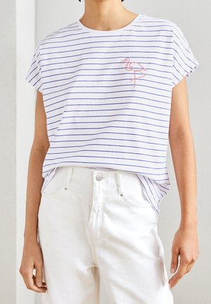 Person iført en hvid kortærmet stribet top med et pink flamingo-design og højtaljede hvide bukser.