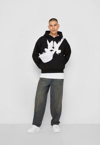 Black hoodie with white bird graphic, layered over white t-shirt. Worn with loose blue jeans and white sneakers, featuring a white crossbody bag.
