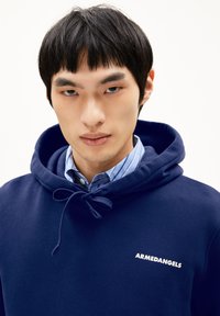 Jonge man met kort zwart haar, gekleed in een blauwe hoodie over een blauw gestreepte shirt, met het gezicht naar voren en een neutrale uitdrukking.