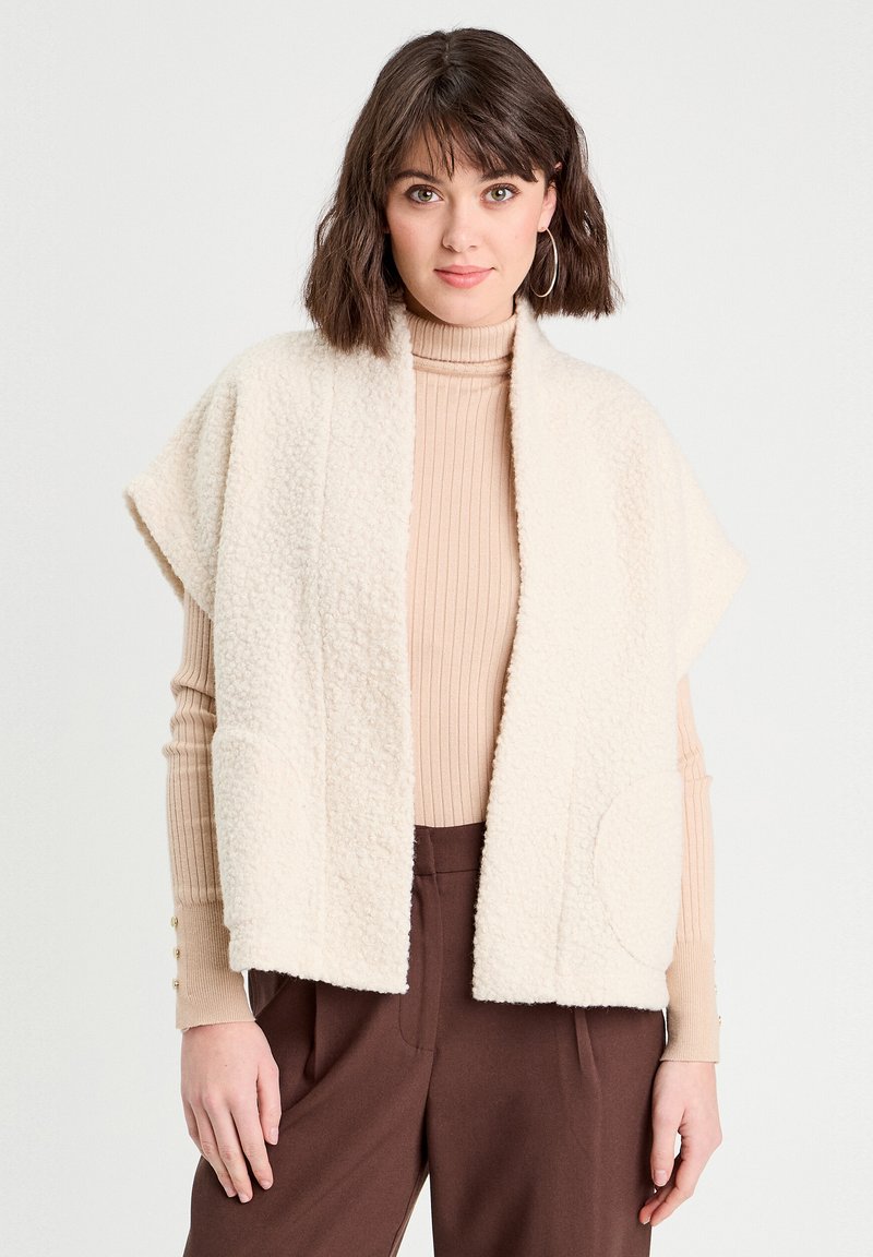 Gilet en polaire couleur crème, avec une ouverture sur le devant, sans manches, et une poche ronde. Porté par-dessus un col roulé beige en côtes et un pantalon marron.