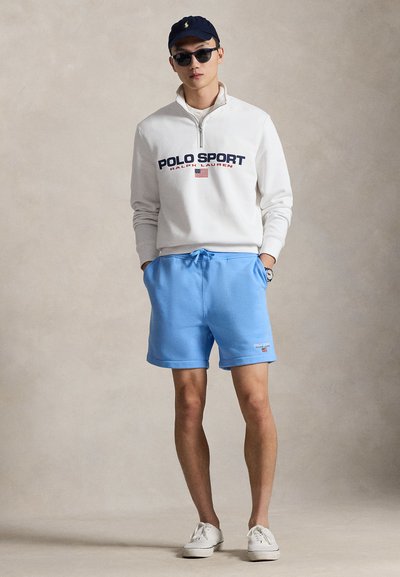 Λευκό φούτερ με φερμουάρ και το κείμενο "POLO SPORT", μπλε σορτς με κορδόνια και λευκά αθλητικά παπούτσια· φορεμένα με γυαλιά ηλίου και σκούφο ναυτικού μπλε.
