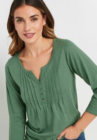 Maglietta verde a manica lunga con scollo rotondo, texture plissè sulla parte anteriore e quattro bottoni sulla chiusura. Tessuto morbido e leggero.