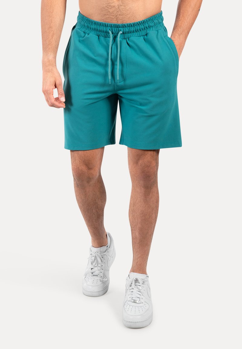 Pantalones cortos de color teal con cintura elástica y cordón. Textura suave y longitud por encima de la rodilla. Combinados con zapatillas blancas.
