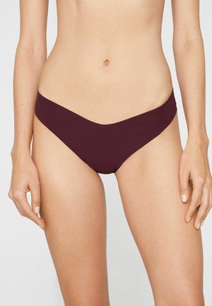 Kobieta ubrana w ciemnofioletową, bezszwową bieliznę bikini z niskim stanem, ukazującą tors od talii do górnej części ud na jednolitym tle.