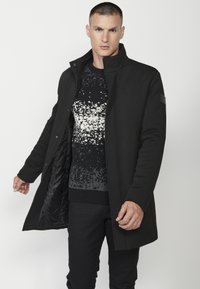 Cappotto nero con colletto alto e zip frontale. Il tessuto appare liscio con una leggera lucentezza. Il modello indossa un maglione in maglia nera a fantasia sotto.