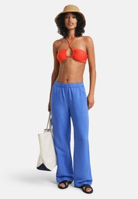 Rød halterneck bikini-topp med et midtsnitt, kombinert med løse blå linbukser og svarte sandaler. En beige stråhatt og en hvit totebag er inkludert.
