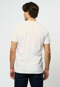 Balta medvilnės marškinėliai su standartiniu kirpimu, trumpomis rankovėmis ir apvaliu iškirpimu, dėvimi asmens, kuris stovi atsisukęs, derinami su tamsiomis džinsinėmis kelnėmis.