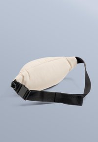 Ljusbeige vadderad ländryggsstödskudde med svart justerbart band och plastspänne. Mjuk textur, oval form och sydda detaljer.