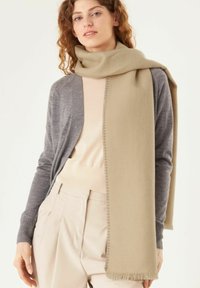 Écharpe beige avec des franges, portée sur un pull léger et un gilet gris. Texture douce, forme rectangulaire et design minimaliste.