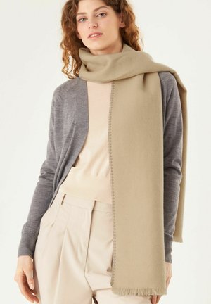 Écharpe beige avec des franges, portée sur un pull léger et un gilet gris. Texture douce, forme rectangulaire et design minimaliste.
