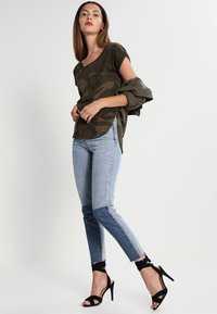 Olive green camo top, loose fit, paired with blue gradient skinny jeans featuring darker patch on lower legs. Black heeled sandals complete the look.