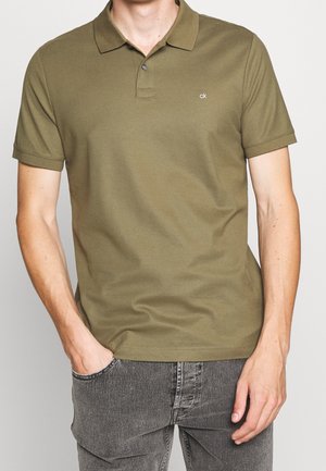 Homme portant un polo manches courtes vert olive avec un petit logo "ck", associé à un jean noir délavé, mains dans les poches.