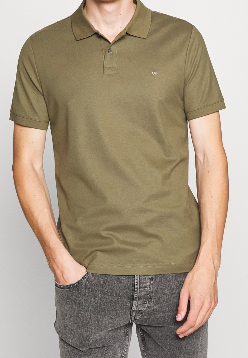 Homme portant un polo manches courtes vert olive avec un petit logo "ck", associé à un jean noir délavé, mains dans les poches.