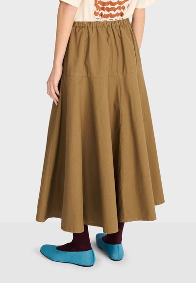 Femme portant une longue jupe marron avec taille élastique, des chaussettes bordeaux foncé et des chaussures plates bleu vif, debout devant un fond uni.