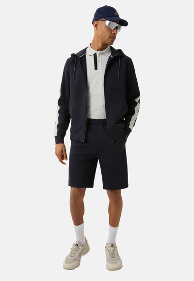 Navy Sportoutfit mit einer Kapuzenjacke mit weißen Streifen, einem grauen Polo mit schwarzem Kragen, passenden Shorts und hellen Sneakers.