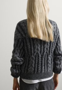Grijze gebreide trui met kabelpatronen, geribde manchetten en zoom; gedragen over een wit shirt. Zachte textuur en chunky ontwerp.