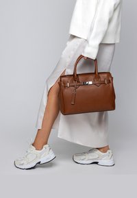 Persoon in witte jurk met zijspleet en sneakers, die een bruine leren handtas aan de handvaten vasthoudt, tegen een effen lichte achtergrond.
