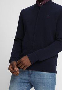 Marinblå zip-up cardigan med en strukturerad mönster, ribbade ärmslut och nederkant. Liten logotyp på bröstet, tillsammans med en rutigt skjorta under.