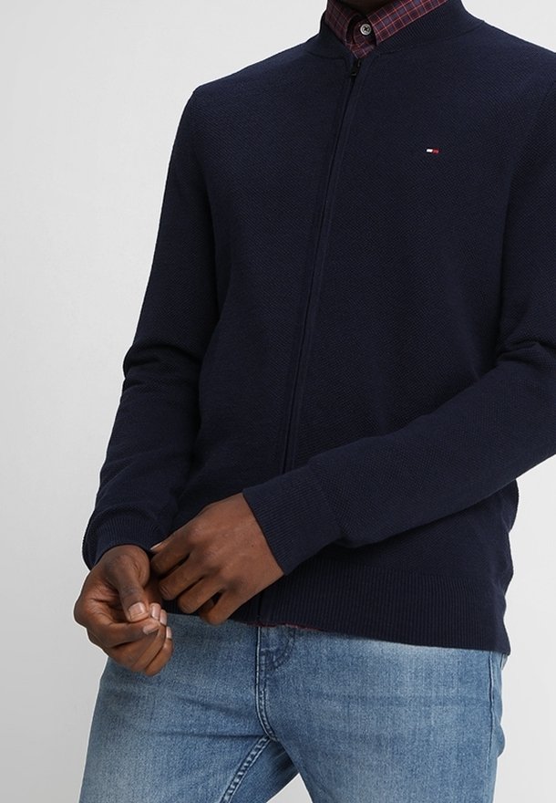 Marinblå zip-up cardigan med en strukturerad mönster, ribbade ärmslut och nederkant. Liten logotyp på bröstet, tillsammans med en rutigt skjorta under.