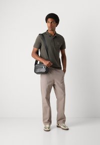Tricou polo verde închis, pantaloni bej deschis și adidași multicolori. Geantă de umăr texturată neagră cu detalii metalice argintii la fermoar.