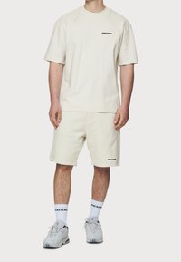 Ljusbeige bomulls t-shirt och shorts-set, med en logotyp på bröstet och benet. Tillsammans med vita strumpor och grå sneakers.