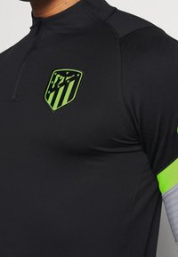 Schwarzes Sportshirt mit halbem Reißverschluss, ausgestattet mit einem lebendigen grünen Logo und Akzenten an den Ärmeln. Das Material wirkt dehnbar und glatt.