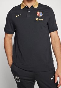 Černé polo tričko se zlatým límečkem, s logem Barcelony a brandingem Spotify. Vyrobeno z texturovaného materiálu; spárováno s tmavými kalhotami.