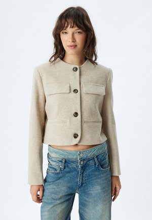 Giacca croppata beige chiaro realizzata in tessuto morbido, con scollo rotondo, quattro bottoni e due tasche sul petto con chiusura a patta.