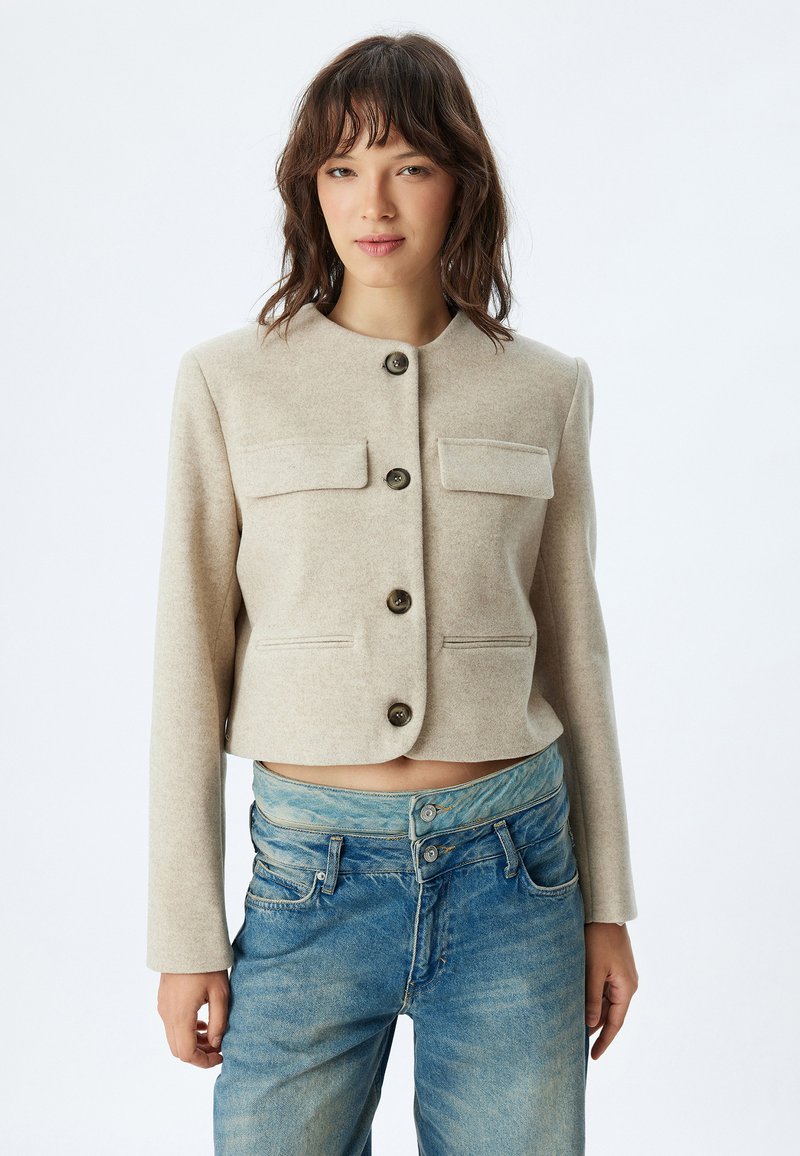 Chaqueta corta de color beige claro, hecha de una tela suave, con cuello redondo, cuatro botones y dos bolsillos en el pecho con solapas.