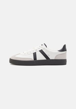 Witte lage sneaker met zwarte strepen, grijze suède accenten op teen en hiel, zwarte zool en witte veters, zijaanzicht op effen achtergrond.