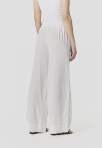 Inimene, kes kannab lahtisi valgeid laia säärega pükse elastse vöökohaga, valget varrukateta topi ja musti sandaale, seisab ühevärvilise tausta ees.