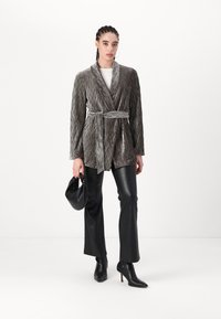 Texturovaný šedý blazer s šálovým límcem a páskem, kombinovaný s černými koženými kalhotami a špičatými kotníkovými botami, držící malou černou tašku.