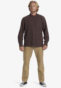 Brown long-sleeve button-up shirt with a chest pocket, paired with beige pants and black sneakers. The shirt has a relaxed fit and textured fabric.
