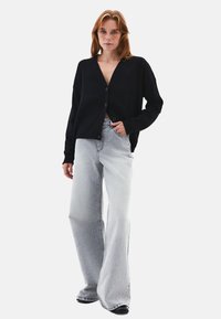 Mustat neuletakki, jossa on V-pääntie ja nappikiinnitys, yhdistettynä vaaleanharmaisiin leveisiin farkkuihin. Kangas näyttää pehmeältä ja siinä on rento istuvuus.