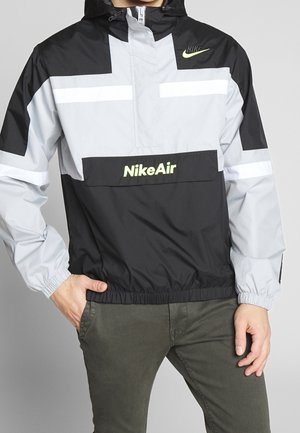 Mężczyzna w czarno-szarej kurtce przeciwwietrznej Nike Air z neonowym logo, stojący z jedną ręką w kieszeni i zielonymi spodniami na szarym tle.
