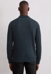Ciemnoturkusowy sweter wykonany z teksturowanej tkaniny, z prążkowanym kołnierzem i dołem. Tył ma luźny krój z minimalną liczbą szwów.
