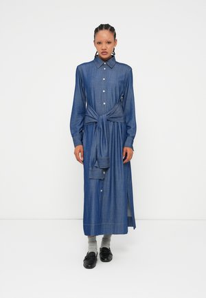 Langdenim jurk met een knoopsluiting aan de voorkant, kraag en een geknoopte taille. Heeft lange mouwen en een zijsplit. Draagt met grijze sokken en zwarte schoenen.