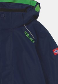 Casaco impermeável azul marinho com detalhes verdes, apresentando o logótipo 'TROLLKIDS', um emblema da bandeira norueguesa e detalhes refletivos ao longo das costuras.