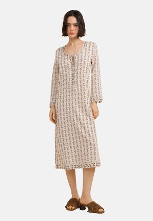 Femme portant une robe beige mi-longue avec des motifs verticaux noirs, manches longues, et des sandales à talons ouvertes marron avec des franges, debout immobile.