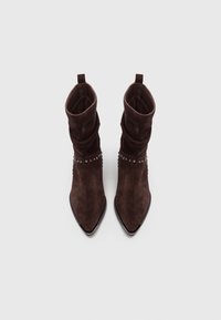 Botas de ante marrón oscuro hasta la mitad de la pantorrilla, con punta afilada, detalles de tachuelas metálicas alrededor del tobillo y un eje arrugado, vistas desde arriba.