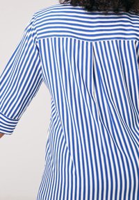 Chemise à rayures bleues et blanches avec un pli au dos, des manches larges à revers, et un design propre et structuré. Tissu doux avec une texture lisse.