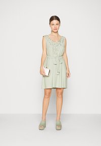 Vestido sem mangas verde claro com bordado floral na gola, fita de tecido na cintura, combinado com sapatos de plataforma verde claro.