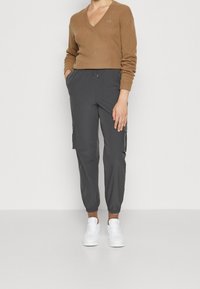 Brun V-ringad tröja i mjukt material, kombinerad med grå cargobyxor med sidofickor, avsmalnande ärmslut och vita sneakers.