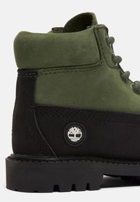 Stivale in pelle alla caviglia di colore verde oliva e nero. Caratterizzato da materiali testurizzati, lacci tondi e una toppa rotonda con logo sul lato. Suola in gomma.