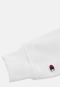 Hvid sweatshirtærme med ribbet manchet, der har et lille broderet logo i rød og sort. Blødt, struktureret stof er synligt.